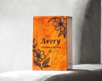 May include: A rectangular wooden cremation urn with a warm orange-brown grain. The front features black laser-engraved floral designs, the name Avery, the dates 1/27/2005 and 3/18/2025, and a small heart symbol centered below the dates.