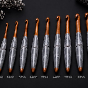Könnte beinhalten: Ein Set aus zehn Häkelnadeln, angeordnet in aufsteigender Größe von 4,0 mm bis 12,0 mm. Jede Nadel hat ein poliertes braunes Holzoberteil mit Haken und einen Griff aus grauem, marmoriertem Harz. Die Größen sind unter jeder Nadel aufgedruckt.