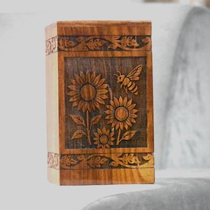 May include: A wooden box with carved floral and bee designs. The front panel features sunflowers and a bee, framed by decorative borders. The box is made of light brown wood and has a rectangular shape.