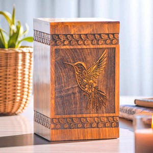 Op de afbeelding: Houten urn met een gebeeldhouwd kolibrie-ontwerp. De rechthoekige doos is gemaakt van warm getint hout, met een gedetailleerde kolibrie-snijwerk op het voorpaneel. Decoratieve bladerpatronen omlijsten de boven- en onderkant.