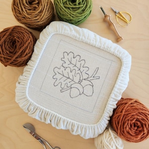 以下が含まれることがあります： オークの葉とドングリがデザインされた白い刺繍枠。枠の周りは、茶色と緑の様々な色の糸の玉で囲まれています。