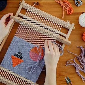 Peut inclure: Un métier à tisser en bois avec un textile partiellement tissé. Le textile présente des motifs géométriques orange, rose et noirs sur fond gris. Divers fils colorés et des ciseaux sont visibles sur la surface en bois.