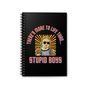 Puede incluir: Cuaderno negro con espiral con el texto "THERE'S MORE TO LIFE THAN... STUPID BOYS" en naranja y azul. La portada presenta una ilustración de una persona con gafas de sol y auriculares.