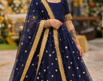 May include: A young girl in a navy blue Indian-style dress with gold accents. The dress features gold polka dots and trim. She is wearing gold earrings and bracelets, and a floral hairpiece. The background is blurred with warm tones.