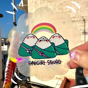 Puede incluir: Una pegatina holográfica con un arcoíris y tres onigiri de dibujos animados, cada uno con una envoltura de alga verde. La pegatina dice "Onigiri Squad" en texto negro.