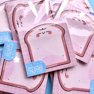 Puede incluir: Un montón de notas adhesivas rosas con un diseño de tostada de dibujos animados. Las notas tienen un patrón de cuadrícula y el texto "sticky notes" está impreso en una de las notas.