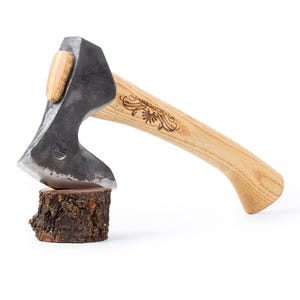May include: A small, hand-forged axe with a wooden handle. The axe head is made of steel and has a slight curve to the blade. The handle is made of light wood and has a decorative design carved into it. The axe is resting on a small piece of wood.