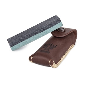 May include: A brown leather case with a stitched design and a small, rectangular sharpening stone inside. The case has a deer head logo on the front.