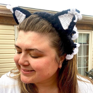 May include: A person wearing a black and white crocheted cat ear headband. The headband has white pom-poms on the ears.