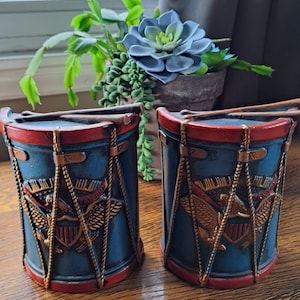 May include: A pair of cylindrical containers shaped like vintage drums, featuring a blue body with red rims and gold-toned rope detailing. Each drum displays a relief emblem of an eagle with a shield and the text Infantry, topped with two drumsticks.