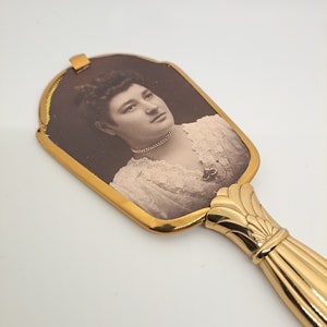 May include: An antique hand mirror with a gold-toned frame and handle. The mirror features a sepia-toned portrait of a woman. The handle has a decorative, fluted design. The mirror is a vintage item.