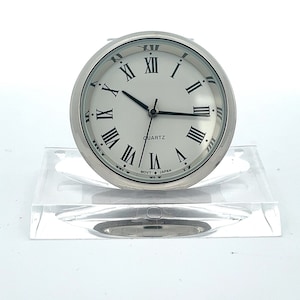 May include: A silver clock with Roman numeral markings on a white face. The clock is on a clear acrylic stand. The clock face reads "QUARTZ MOV'T JAPAN".