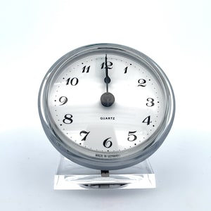 Puede incluir: Un reloj plateado con una esfera blanca y números negros. El reloj está puesto a las 12:00 y tiene las palabras "QUARTZ" y "MADE IN GERMANY" impresas en la esfera.