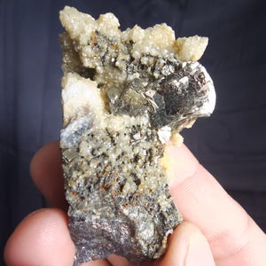 May include: A natural mineral specimen featuring a cluster of pyrite crystals with a metallic luster. The specimen has a dark gray and silver color with areas of beige and white crystalline formations. The rough texture and unique shape highlight its raw, natural beauty.