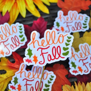 May include: Five white stickers with the text "hello fall" in orange, red, and green lettering. The stickers are surrounded by autumn-coloured leaves.
