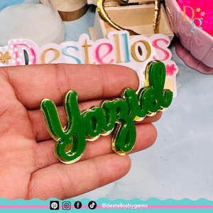 Puede incluir: Un letrero de nombre de acrílico verde con ribete dorado, deletreando el nombre "Yaziel". El letrero se sostiene en una mano.