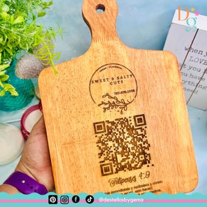Puede incluir: Una tabla de cortar de madera con un asa, con el texto "SWEET & SALTY CUTS" y un código QR. La tabla es de color marrón claro y tiene vetas de madera natural. El texto "Filipenses 4:9" también es visible.
