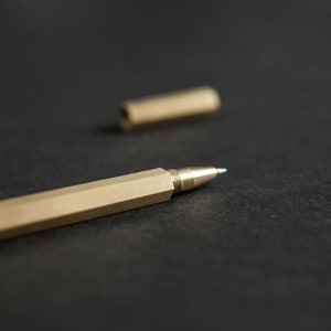 May include: A gold-coloured pen with a hexagonal barrel and a silver tip. The pen is lying on a black surface.