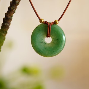 May include: A jade donut pendant necklace. The green stone is round with a hole in the center, suspended from a brown cord. Gold-colored beads accent the cord. The background is blurred, with a tree branch in the upper left.