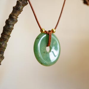 May include: A green jade donut pendant necklace with a brown leather cord. The pendant is a light green color with a hole in the center. The necklace is hanging from a tree branch.