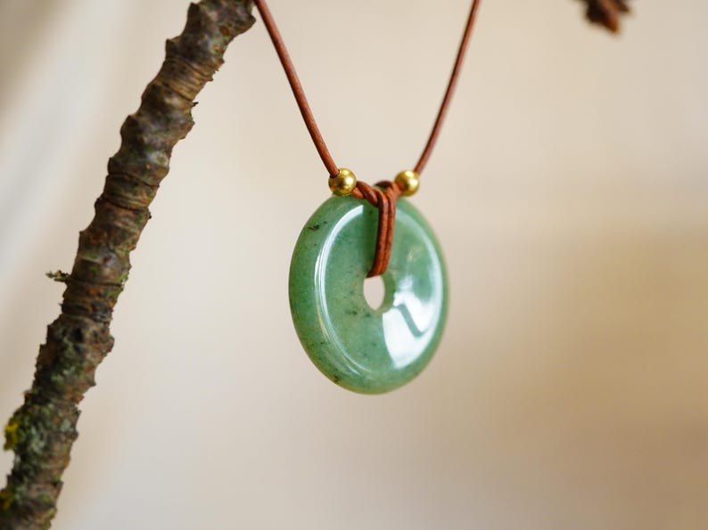May include: A green jade donut pendant necklace with a brown leather cord. The pendant is a light green color with a hole in the center. The necklace is hanging from a tree branch.