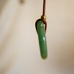 May include: A jade pendant with a polished, elongated shape. The green stone is suspended from a brown cord with a gold-colored bead. The pendant is a simple, elegant piece of jewelry.