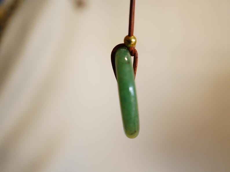 May include: A jade pendant with a polished, elongated shape. The green stone is suspended from a brown cord with a gold-colored bead. The pendant is a simple, elegant piece of jewelry.