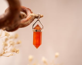 May include: A carnelian crystal pendant with a silver-colored bail, suspended from a curved, brown branch. The pendant is a vibrant orange-red color and is faceted into a hexagonal shape. The bail is stamped with "925".