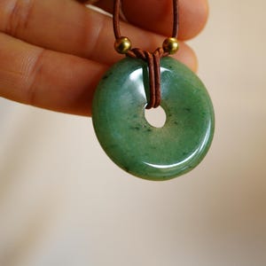 May include: A green jade donut pendant necklace. The polished stone has a central hole and is strung on a brown leather cord with gold-colored beads. The pendant is held up by a hand.