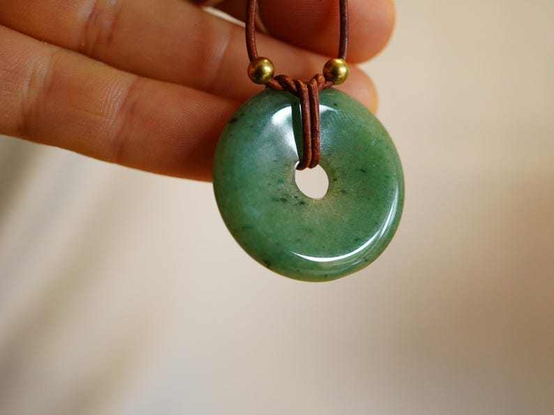 May include: A green jade donut pendant necklace. The polished stone has a central hole and is strung on a brown leather cord with gold-colored beads. The pendant is held up by a hand.