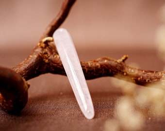 May include: A smooth, pale pink crystal wand, approximately 5 inches long, rests against a brown, textured branch. The wand has a rounded tip and a polished surface, reflecting soft light. The background is a blurred, warm-toned setting.