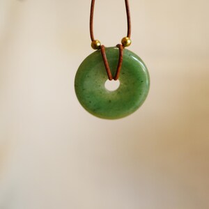 May include: A green jade donut pendant necklace. The circular pendant is a light green color with a hole in the center. The pendant hangs from a brown leather cord with gold-colored beads. The background is a soft, neutral color.
