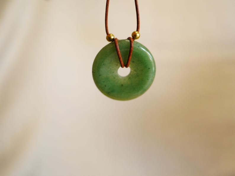 May include: A green jade donut pendant necklace. The circular pendant is a light green color with a hole in the center. The pendant hangs from a brown leather cord with gold-colored beads. The background is a soft, neutral color.