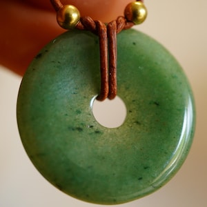 May include: A green jade pendant with a circular shape and a central hole. The pendant is strung on a brown leather cord, with gold-colored beads at the top. The jade has a polished, glossy finish.