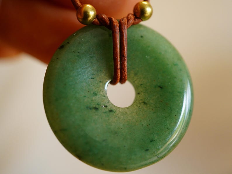 May include: A green jade pendant with a circular shape and a central hole. The pendant is strung on a brown leather cord, with gold-colored beads at the top. The jade has a polished, glossy finish.