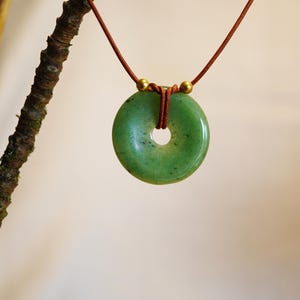 May include: A green jade donut pendant necklace. The circular pendant has a hole in the center and is suspended from a brown cord. Two gold-colored beads are at the top of the pendant. The necklace is displayed against a neutral background.