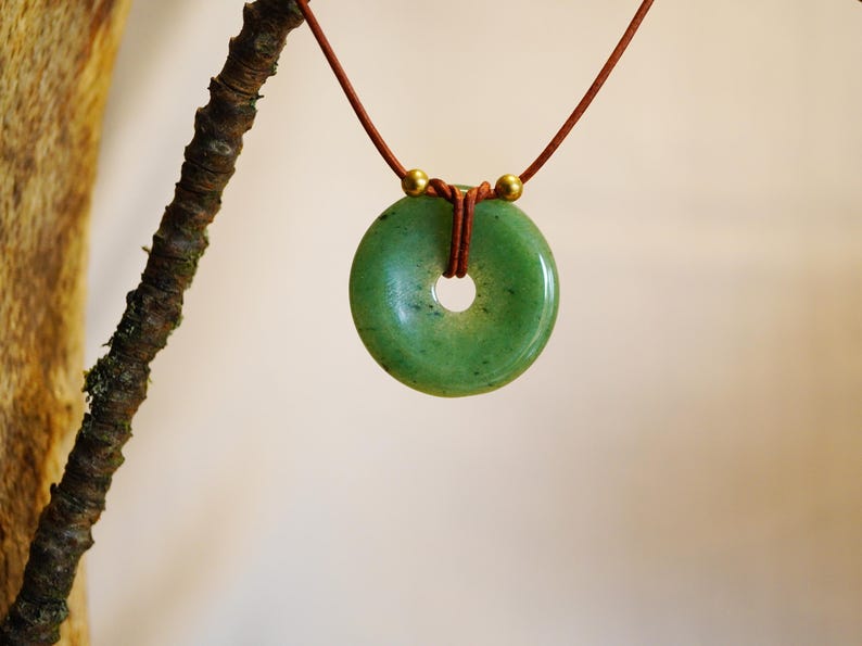 May include: A green jade donut pendant necklace. The circular pendant has a hole in the center and is suspended from a brown cord. Two gold-colored beads are at the top of the pendant. The necklace is displayed against a neutral background.