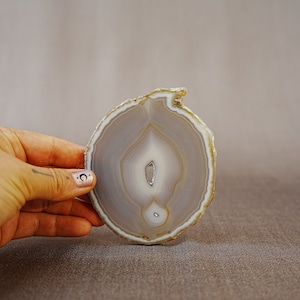 May include: A light grey agate slice with a white centre and brown edges. The agate is translucent and has a natural, organic shape.