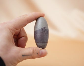 May include: A smooth, oval-shaped stone with a gray and purple color gradient. The stone is held between fingers, showcasing its polished surface and natural variations. The object is a decorative item.