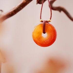 Könnte beinhalten: Ein roter und orangefarbener Achat-Anhänger an einer braunen Lederschnur. Der Anhänger ist donutförmig mit einem Loch in der Mitte.