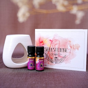 Könnte beinhalten: Zwei kleine Glasflaschen mit ätherischem Öl mit Etiketten, auf denen "Orange & Ylang Ylang" und "Rose & Mimosa" steht. Die Flaschen stehen auf einem weißen Keramikdiffusor mit einer kleinen Öffnung in der Mitte. Hinter den Flaschen befindet sich eine weiße Karte mit einem Aquarellhintergrund und dem Wort "SELBSTLIEBE" in schwarzer Schrift.