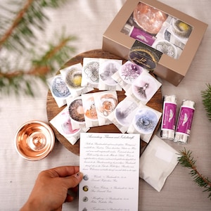 Könnte beinhalten: Eine Auswahl an Produkten mit floralem Thema, darunter kleine, runde Verpackungen mit verschiedenen Designs, eine Kupferschale und zwei zylindrische Artikel mit lila Etiketten. Eine Schachtel mit ähnlichen Artikeln ist sichtbar.