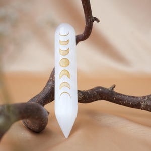 Könnte beinhalten: Ein weißer Selenitstab mit eingeätzten goldenen Mondphasen. Der Stab wird vor einem braunen Ast und einem weichen, neutral getönten Hintergrund präsentiert. Der Stab ist etwa 15 cm lang.