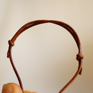 May include: A close-up of a brown leather bracelet with a knotted closure. The bracelet is made of two thin, parallel leather cords that form a loop. The background is a soft, neutral color.