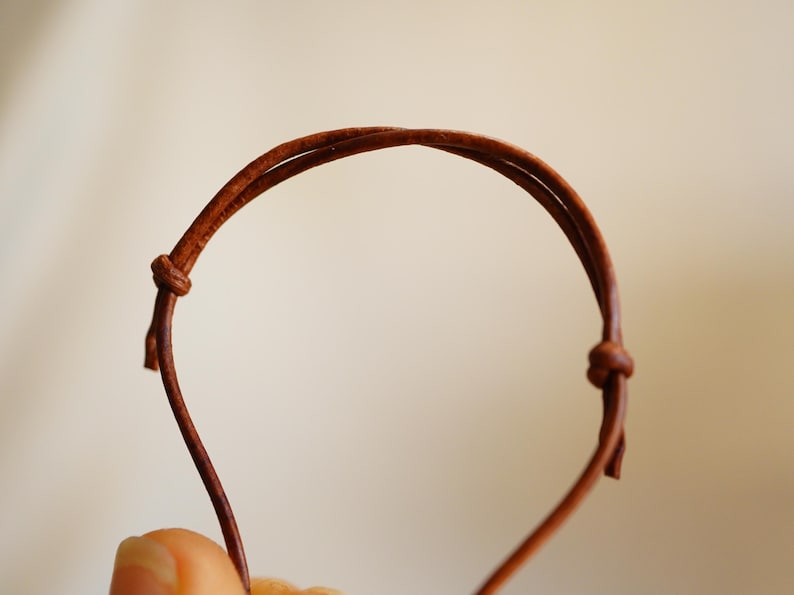May include: A close-up of a brown leather bracelet with a knotted closure. The bracelet is made of two thin, parallel leather cords that form a loop. The background is a soft, neutral color.