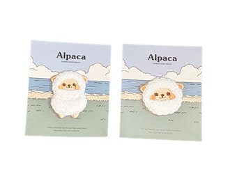May include: Two white cards with a cartoon alpaca on each. The alpaca is white with a brown face and a pink nose. The cards have a blue and green background with a beach scene. The text on the cards reads "Alpaca Embroidery Patch".