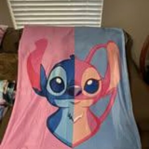 Może przedstawiać: Koc z polaru z podzielonym wzorem z postaciami Stitch i Angel z filmu animowanego Lilo & Stitch. Lewa strona jest r&oacute;żowa ze Stitchem, a prawa strona jest niebieska z Angel.