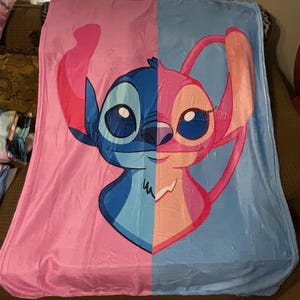 Może przedstawiać: Duży, miękki koc z podzielonym wzorem przedstawiającym postacie Stitcha i Angel z serii Lilo & Stitch. Lewa strona jest r&oacute;żowa, a prawa niebieska. Koc wykonany jest z pluszowego materiału.