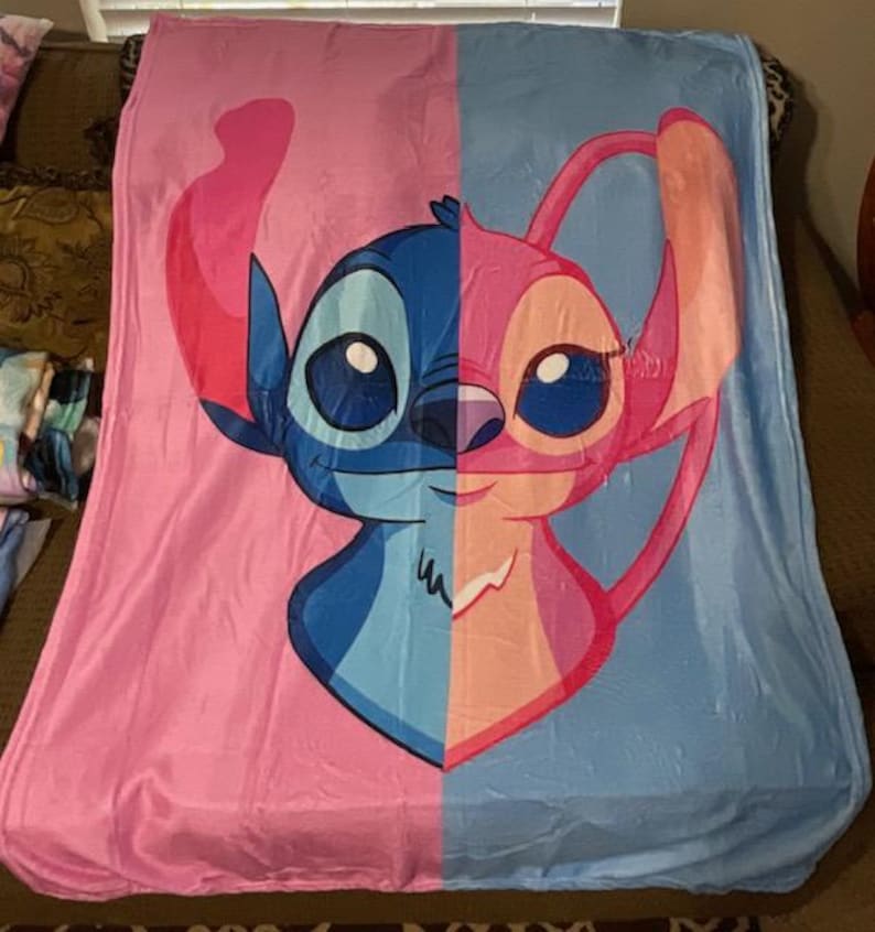 Może przedstawiać: Duży, miękki koc z podzielonym wzorem przedstawiającym postacie Stitcha i Angel z serii Lilo & Stitch. Lewa strona jest r&oacute;żowa, a prawa niebieska. Koc wykonany jest z pluszowego materiału.