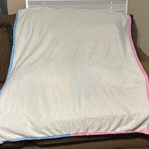 Może przedstawiać: Miękki, biały koc z niebieską i r&oacute;żową obw&oacute;dką jest narzucony na brązową sofę. Koc ma miękką, pluszową fakturę i wydaje się być wykonany z przytulnego materiału. Krawędzie są starannie wykończone kontrastową lam&oacute;wką.